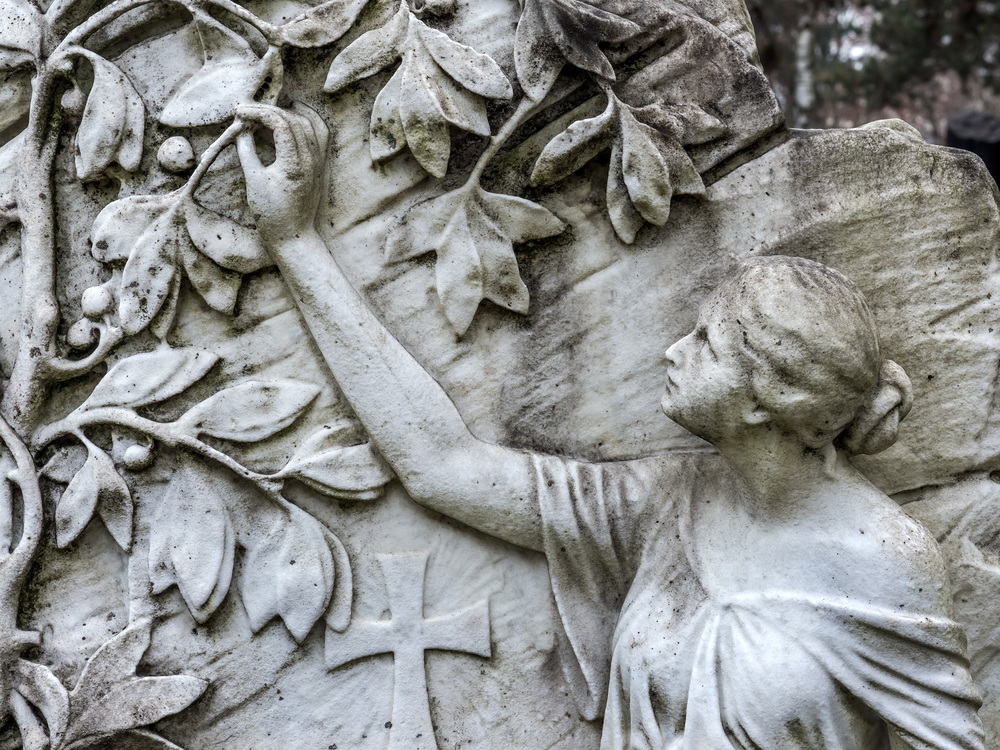 Grabstein auf einem Friedhof mit Engel der an einem Baum Obst pflückt