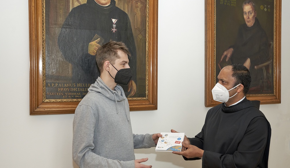 Pater Provinzial Saji Mullankuzhy überreicht in der Wiener Provinzverwaltung der Barmherzigen Brüder ein Exemplar des Buches „Kleine Gase – Große Wirkung: Der Klimawandel“ an einen Mitarbeitenden.