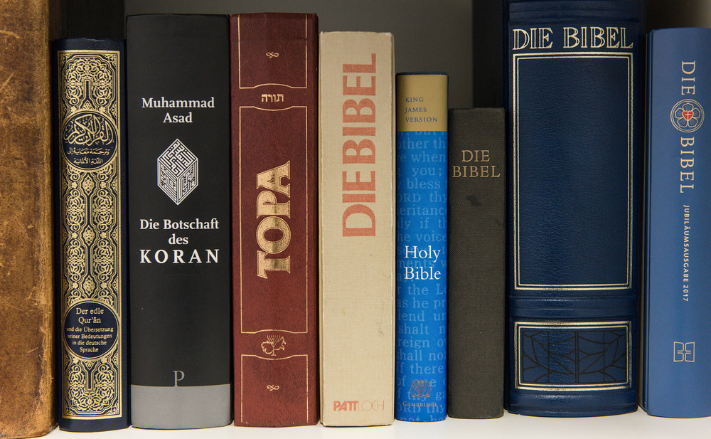 Blick auf die Buchrücken verschiedener Ausgaben des Koran, der Thora und der Bibel nebeneinander in einem Bücherregal. Die Bücher sind die heiligen Schriften der drei abrahamitischen Religionen Islam, Judentum und Christentum.