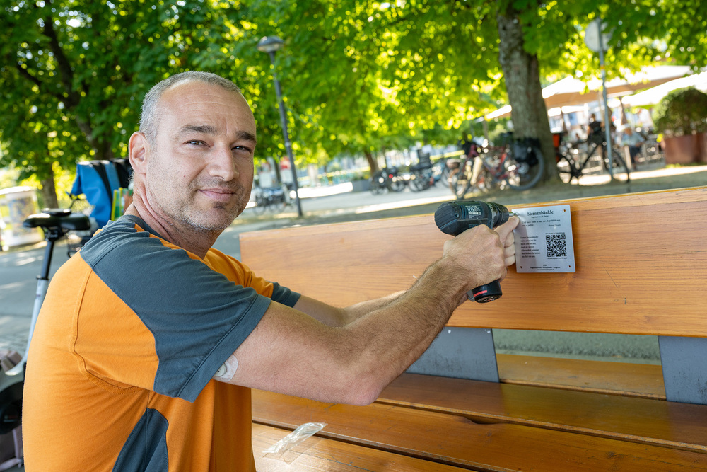 Mann schraubt Plakette an Bank / Alexandra Serra / PK Sternenbänkle Vorarlberg: 'Sternenbänkle' erinnern an früh verstorbene Kinder